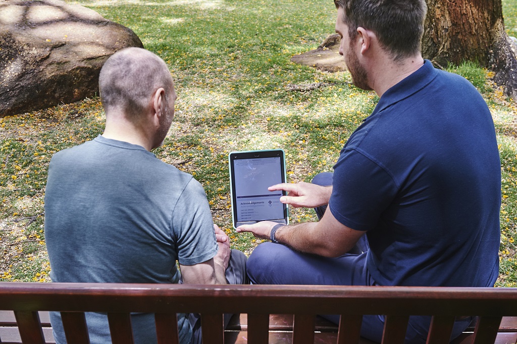 Two men seated on a bench, engaged with a tablet computer, discussing insights from cancer case studies.